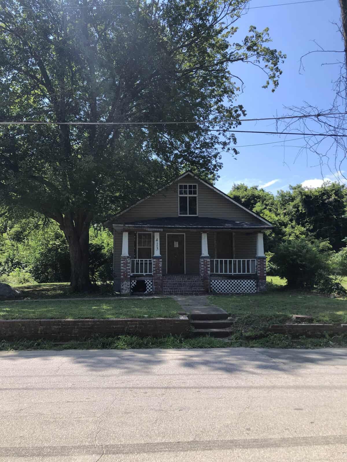 A small, two-story house with a gabled roof and a front porch, located on a street with a sidewalk in the foreground, surrounded by greenery on a sunny day.