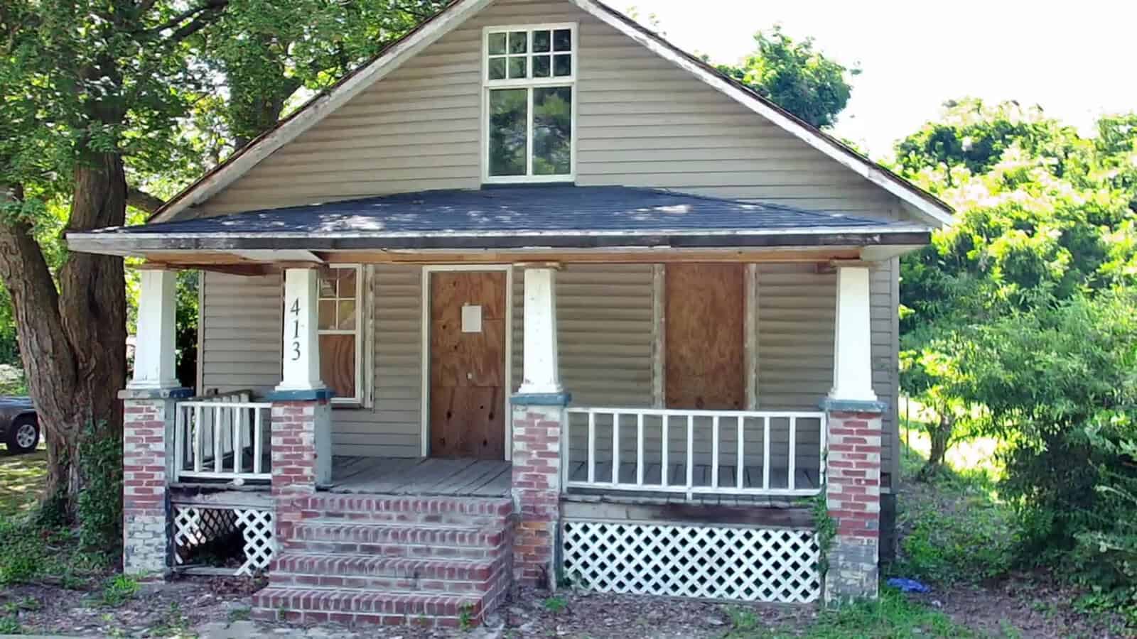 A small, one-story house with a covered front porch and boarded-up windows and door.