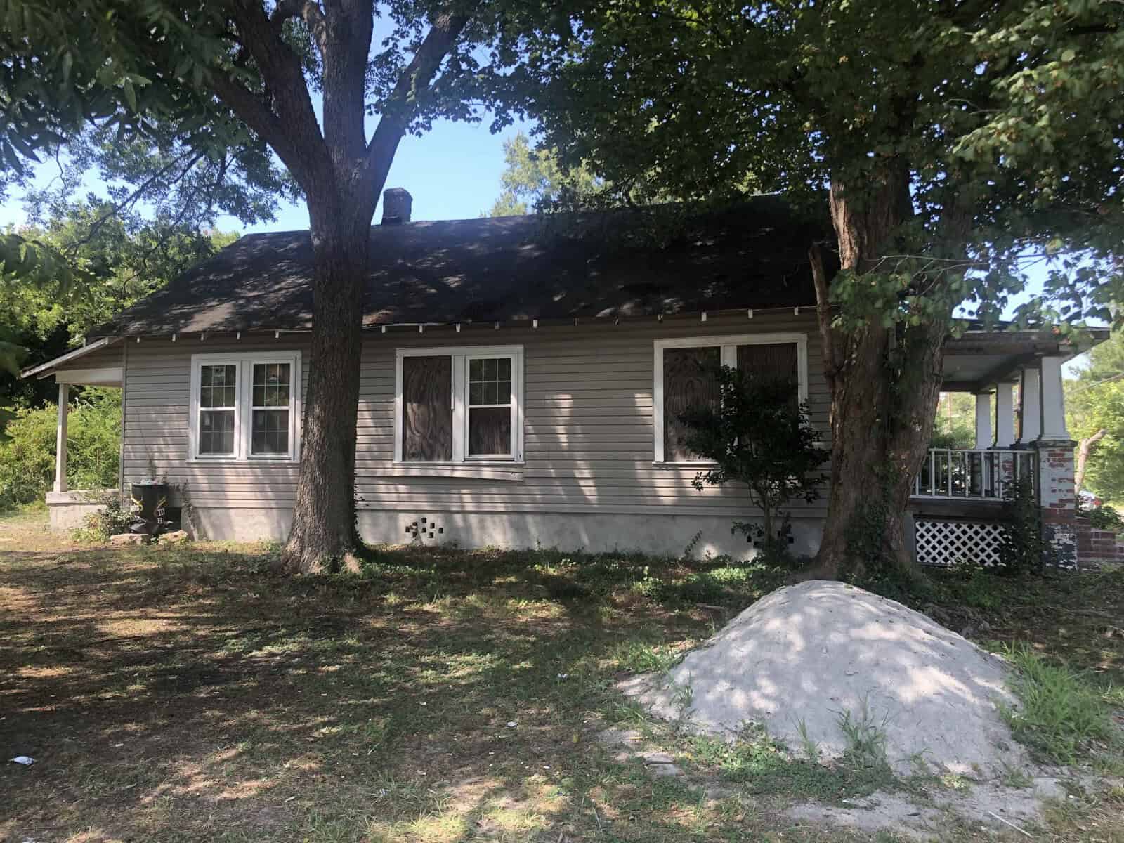 A single-story older house with a sloping roof and weathered gray siding sits under a large tree, with a dirt yard and some foliage around.