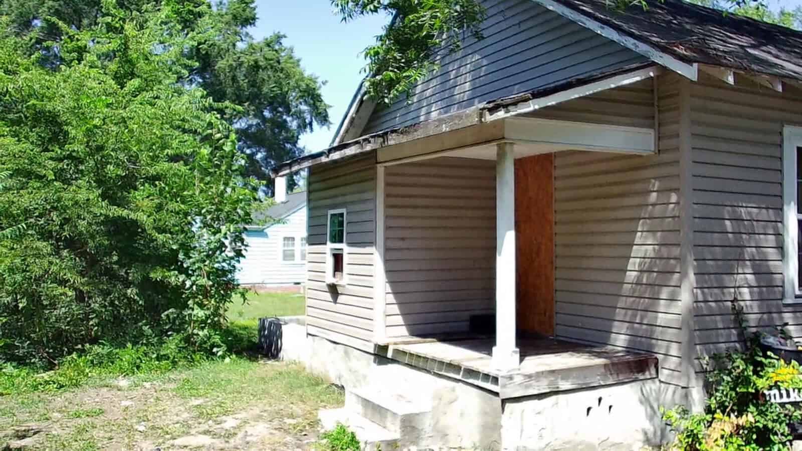 A weathered, small, wooden house with boarded-up windows, situated next to an overgrown yard, under a clear sky.