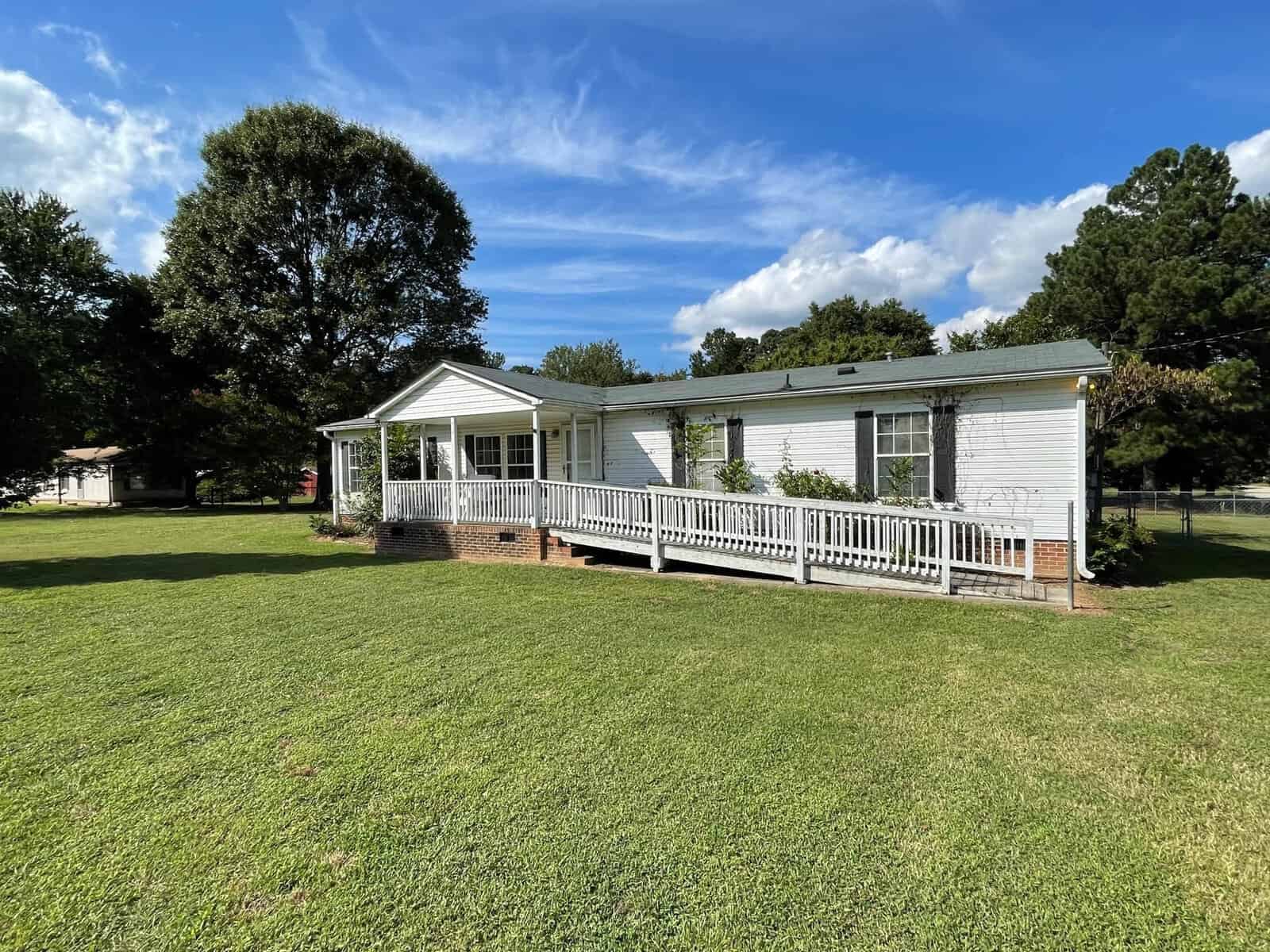A white single-story house with a large front porch and ramp, featuring a well-maintained lawn under a clear sky with scattered clouds.
