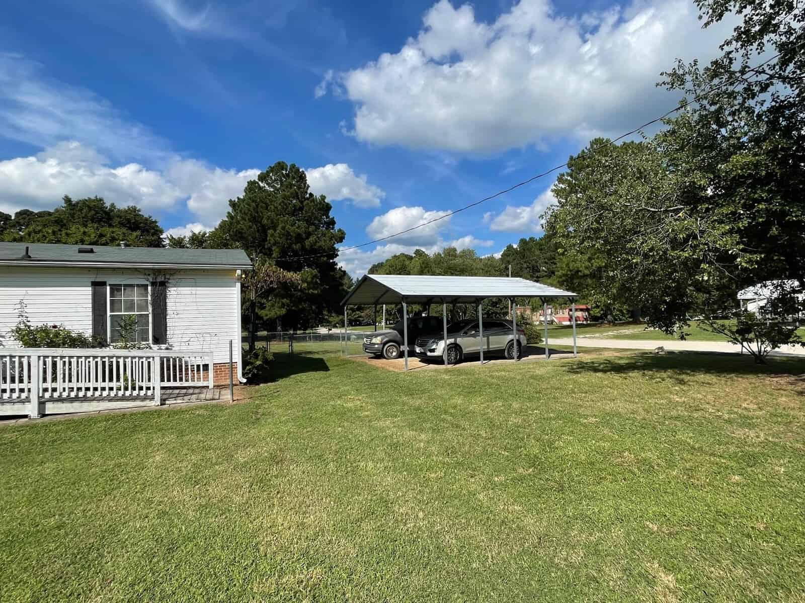 A white single-story house with a covered front porch and a carport sheltering two golf carts, situated on a lawn with trees in the background under a partly cloudy sky.