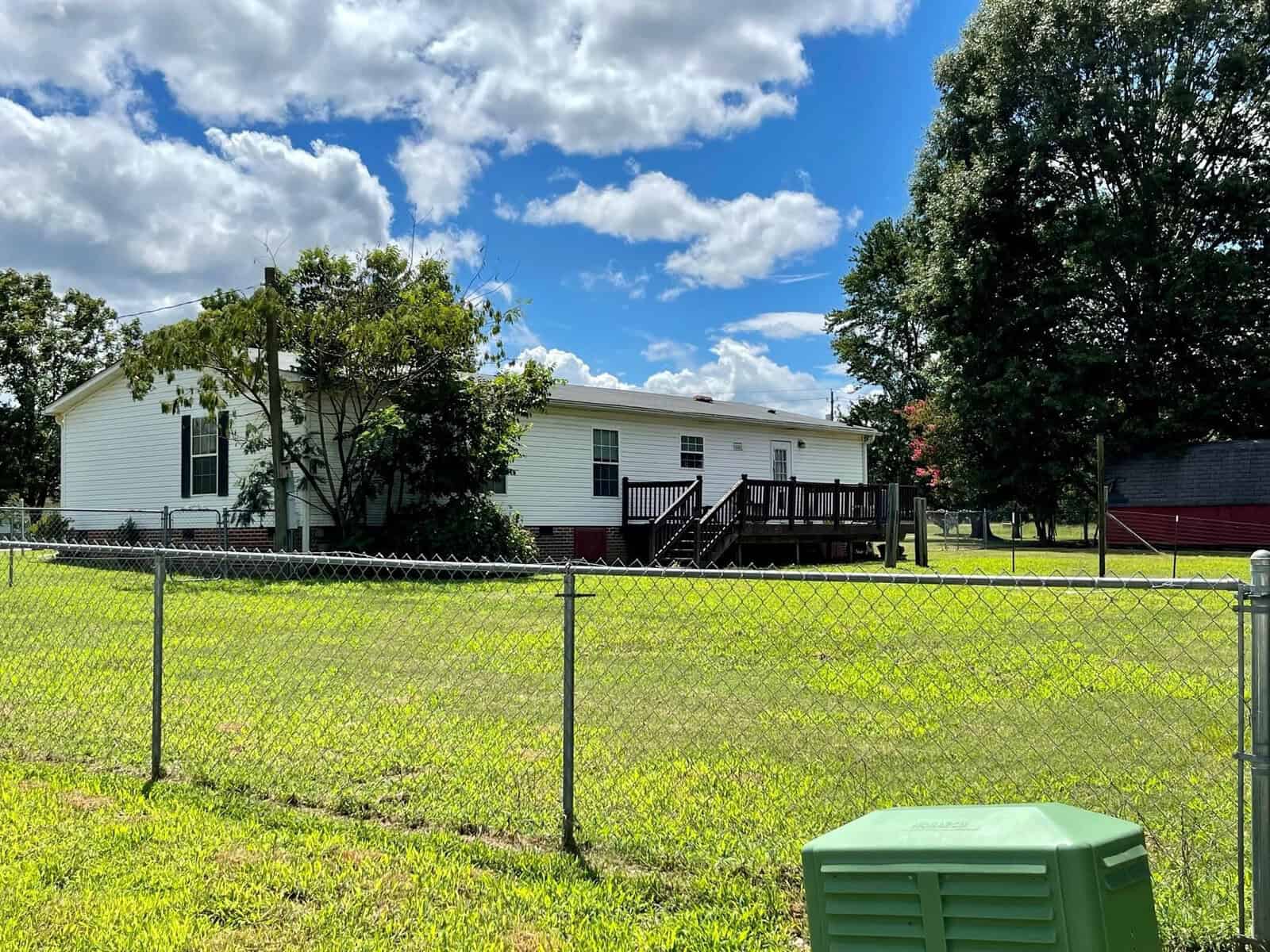 A white mobile home with a black-stairs deck behind a chain-link fence, situated in a grassy area with a few trees under a partly cloudy sky.
