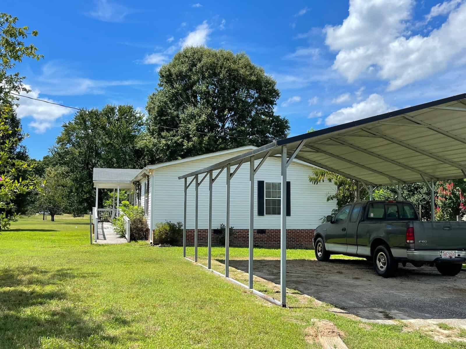 A single-story white house with a metal carport sheltering a pickup truck, set against a backdrop of greenery on a sunny day.