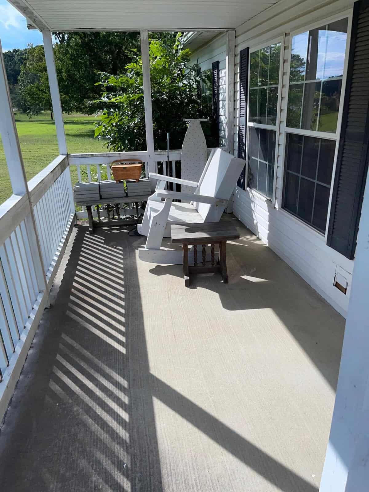 A sunlit porch with white railings featuring an arrangement of outdoor furniture, including a white adirondack chair and a small wooden table, with a view of the lawn and trees.
