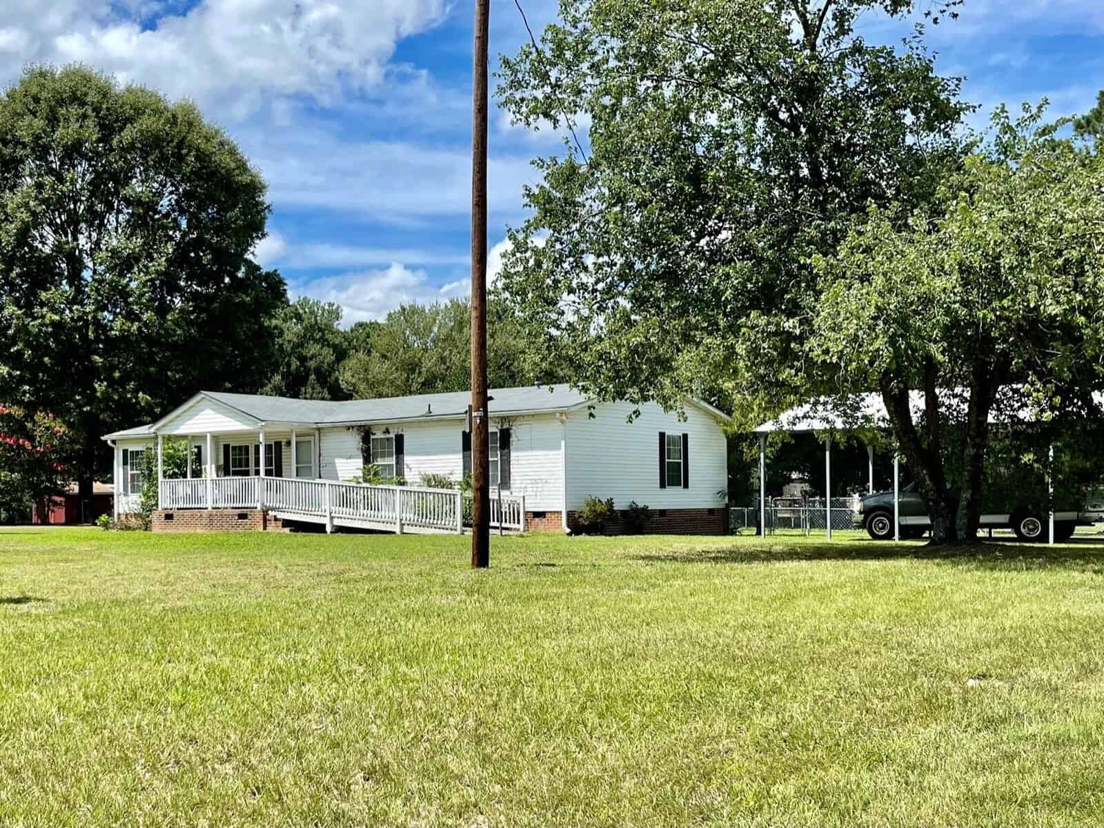 A single-story white house with an attached covered deck, surrounded by a green lawn and trees, under a blue sky with scattered clouds.