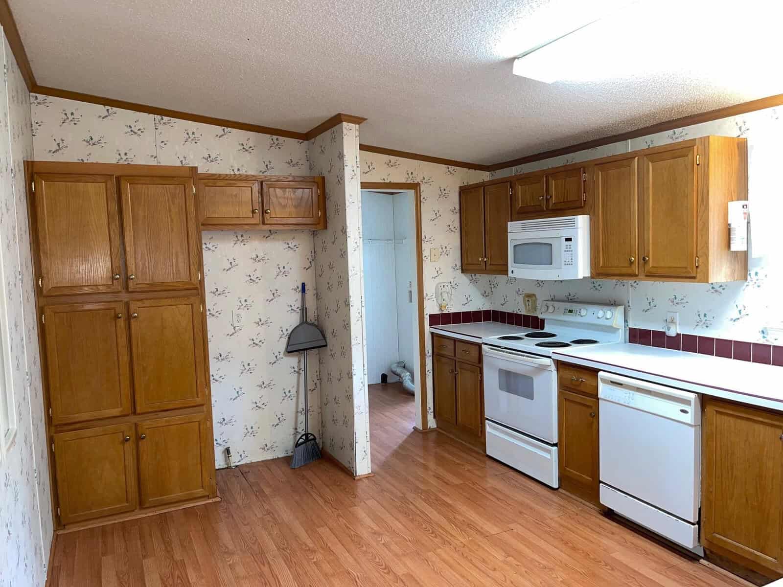A dated kitchen with wooden cabinets, white appliances, and floral wallpaper.