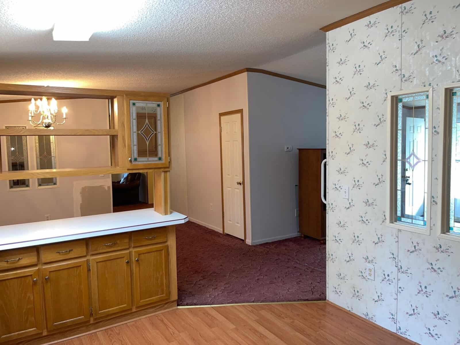 An interior view of a room featuring wooden cabinetry and counters, a floral wallpapered wall with a mounted mirror, a chandelier, and carpeted flooring leading into an adjoining room with a closed white door.