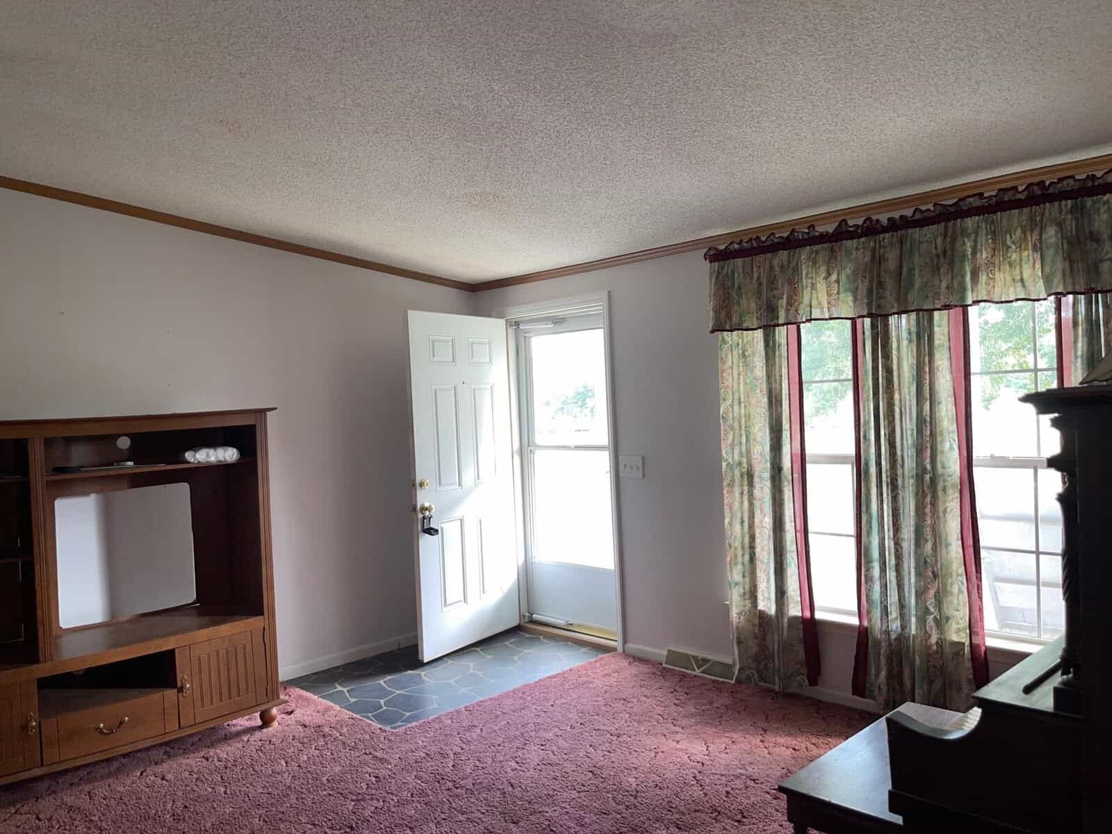 An empty room with a white door, pink carpet, a window with patterned curtains, and some wooden furniture.