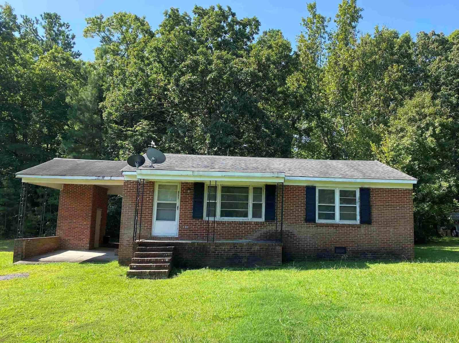 Nestled at 2801 Burnside Road, this charming red brick house in Oxford features a carport and satellite dishes on the roof. A few steps lead to the front door, flanked by shutters on two windows. Lush green grass surrounds it, with trees standing majestically in the background.