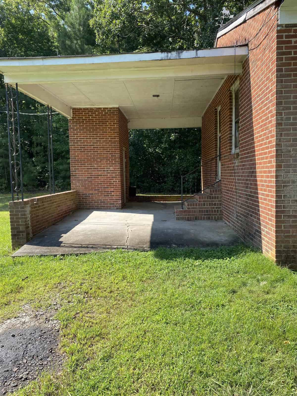 A brick building with a covered porch and concrete steps leading to a doorway. The area is surrounded by green grass and there are trees in the background. The porch features black metal support columns.