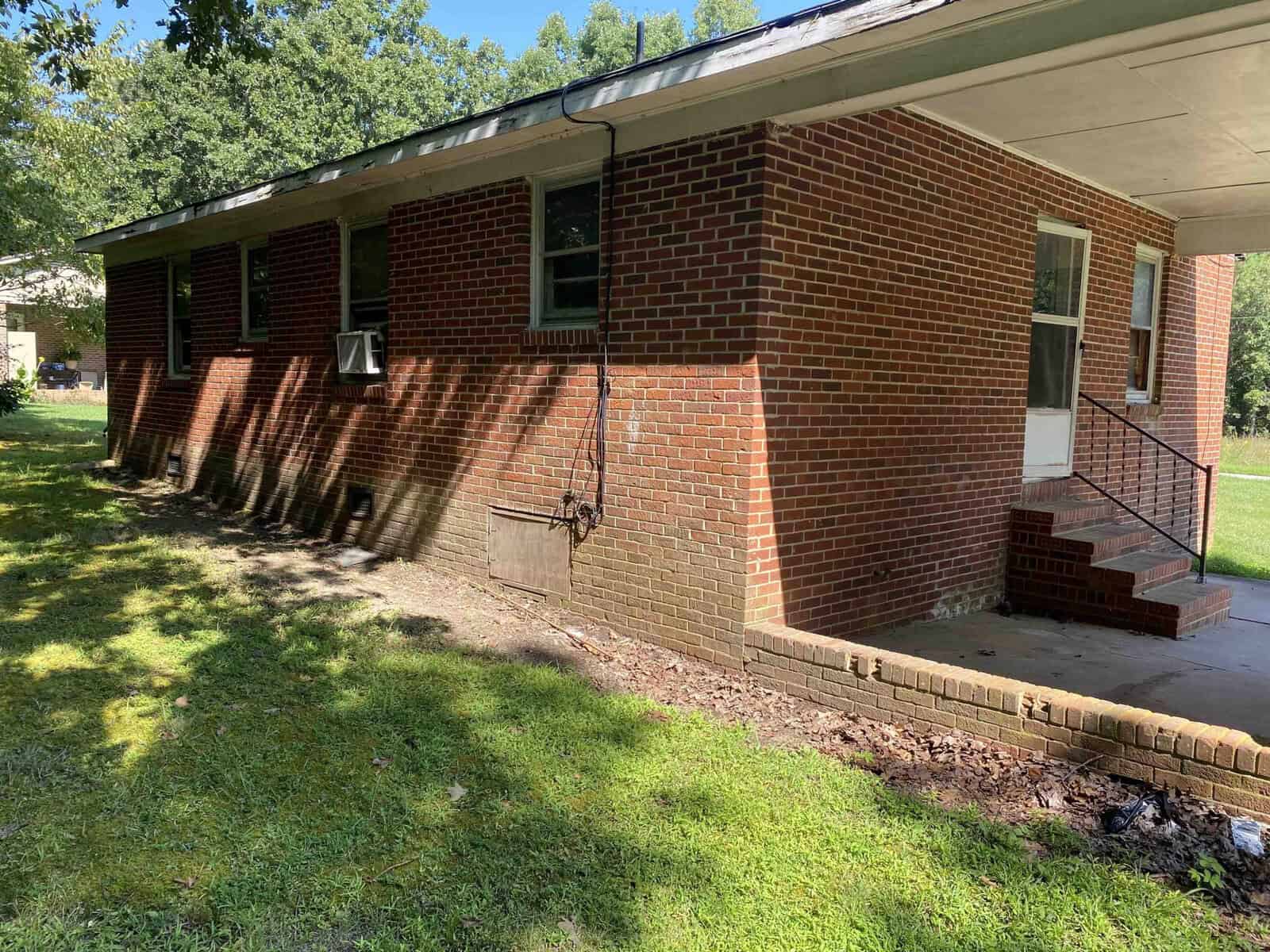 A brick house with a small porch, steps leading to a white door, and a window air conditioning unit. The building is surrounded by grass and shaded by trees, creating a peaceful, suburban atmosphere.