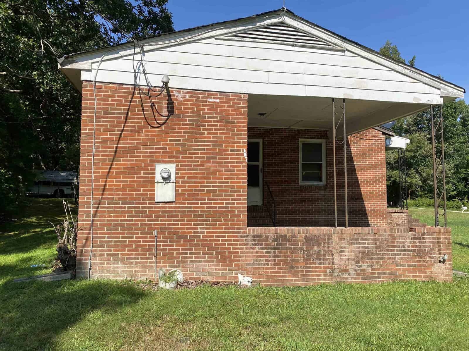 A small brick house with a covered front porch and grass lawn. The house has a white gable above the porch and cables attached to the side. The front yard is green and sunny, with trees in the background.