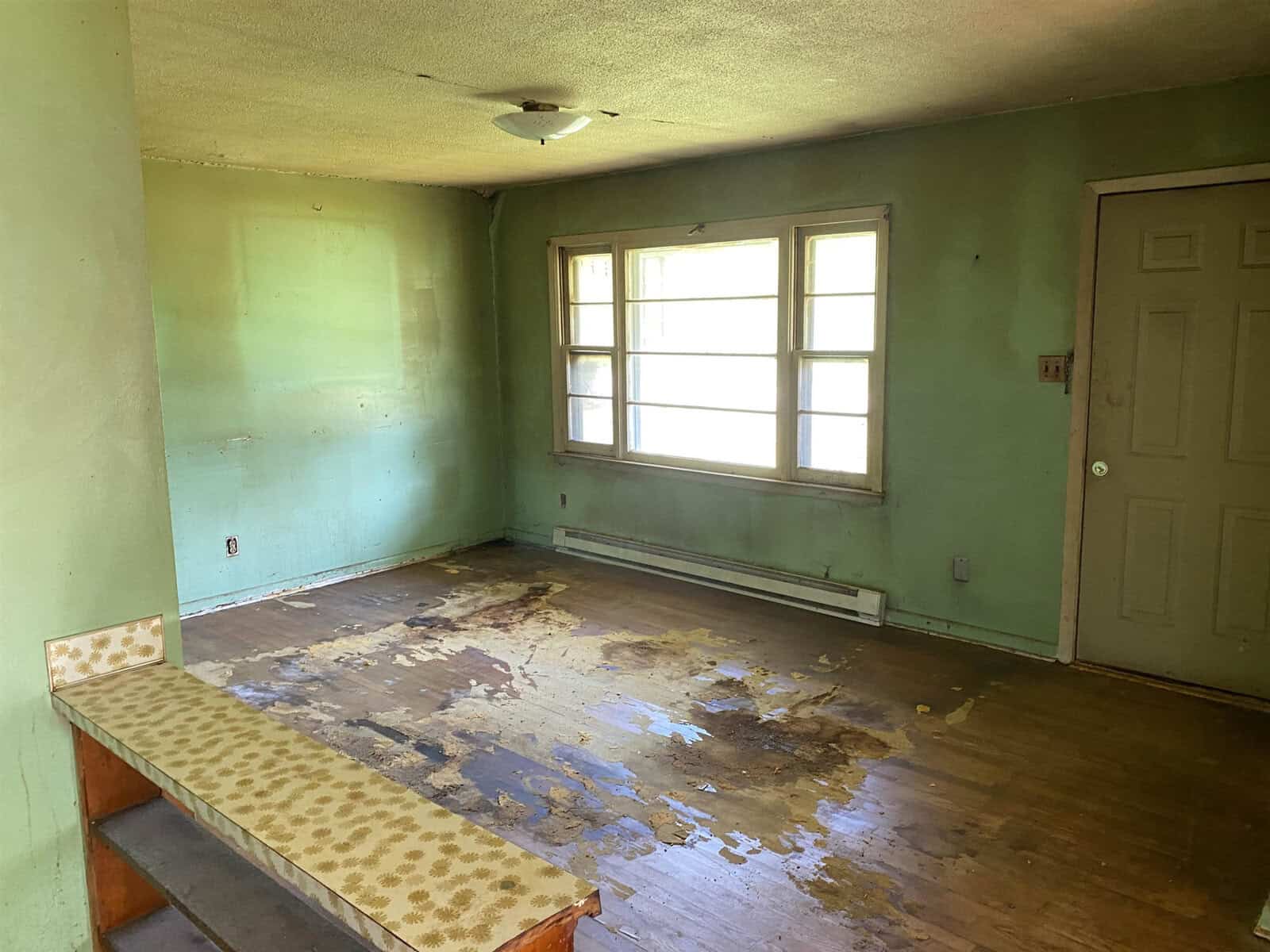 A small room with green walls and a wooden floor showing significant water damage. There's a large window next to a closed door, and a built-in ledge with patterned wallpaper on the left side. The ceiling has a light fixture.