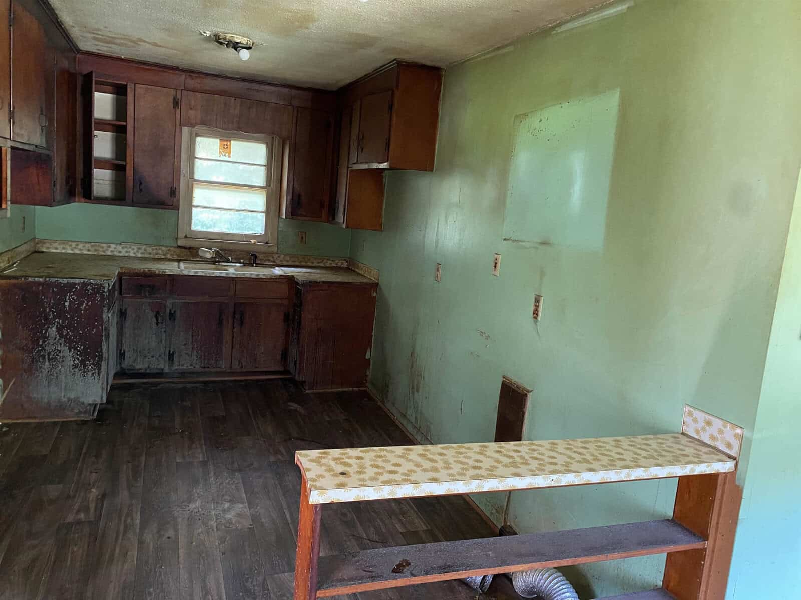 A dilapidated kitchen with peeling paint on green walls. The wooden cabinets and countertop are worn and water-damaged. An old light fixture hangs from the ceiling, and a bench with a patterned cushion is in the foreground. The floor appears dark and stained.