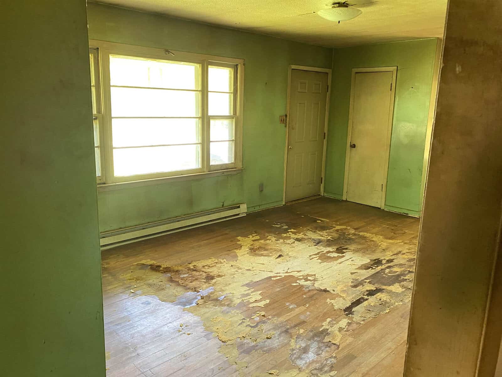 An empty room with worn wooden floors and peeling paint. The walls are green, with a large window letting in natural light. A white door is on the right, and there's visible damage on the floorboards. The ceiling has a small, simple light fixture.