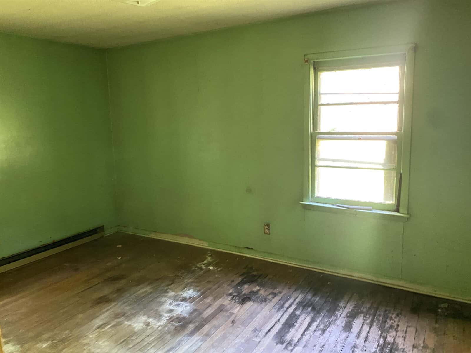 A dimly lit room with green walls and a wooden floor, showing signs of wear and water damage. A single window allows light to enter, casting a soft glow on the weathered surfaces.