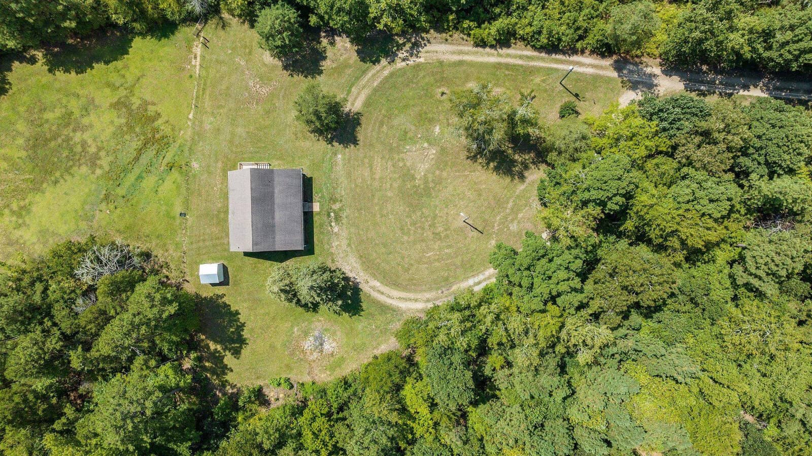 Aerial view of a solitary building with a gray roof surrounded by green fields and trees, with a curved dirt path leading around it.