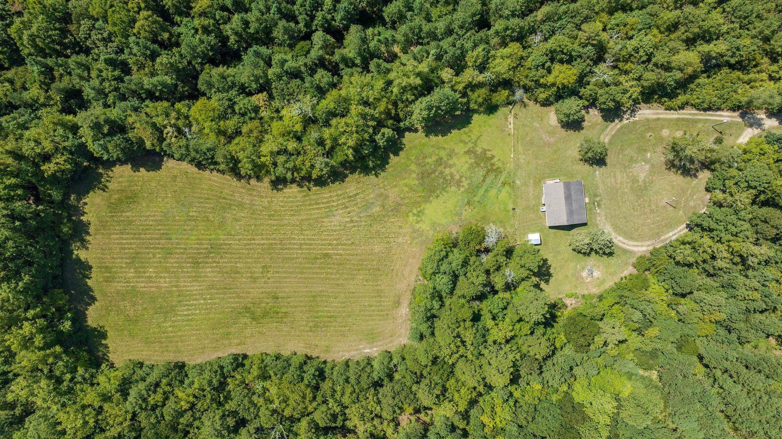 Aerial view of a rural landscape with a mowed field, a small building with a white roof, surrounded by dense trees.