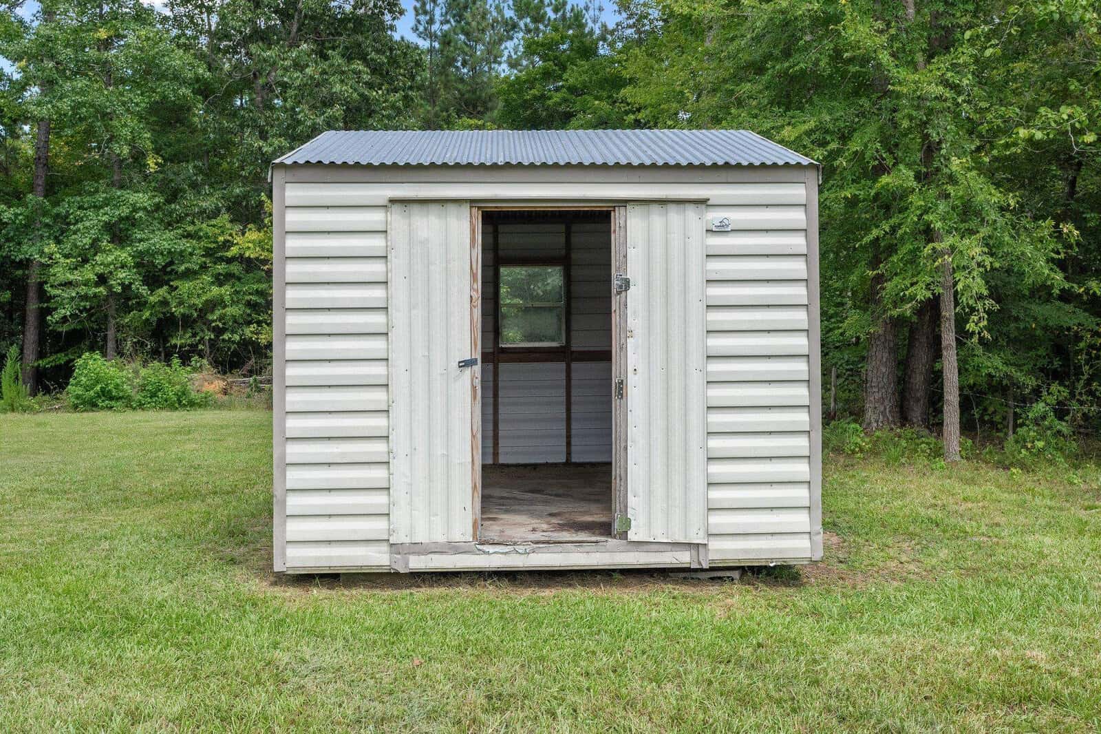 A small, white metal shed with an open door situated in a grassy area surrounded by trees.