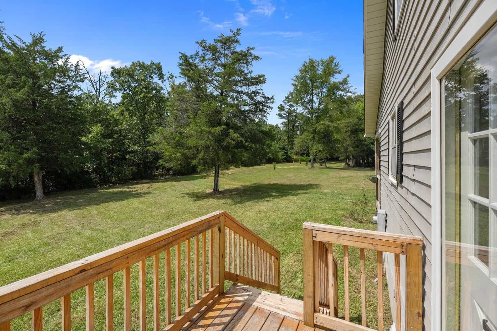 Wooden deck of a house overlooking a spacious backyard with green grass and scattered trees under a clear blue sky.