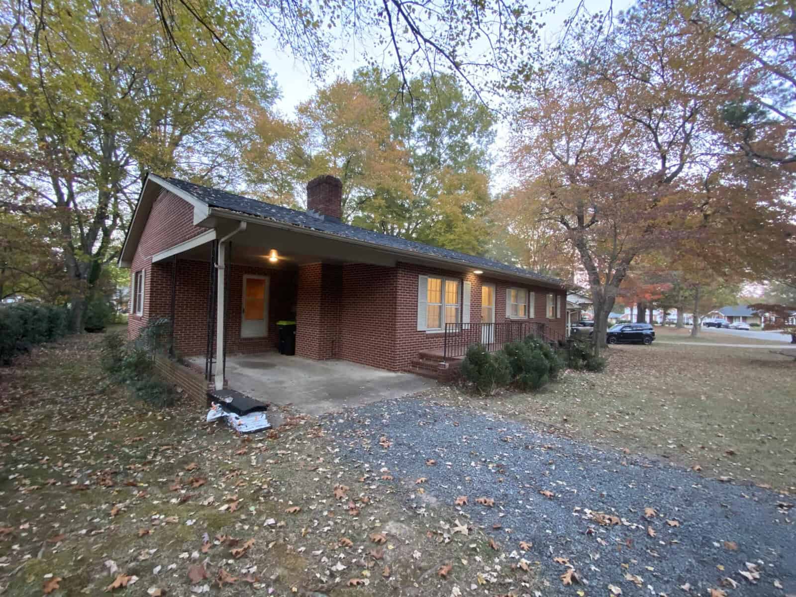 A brick house with a covered porch surrounded by autumn trees. Fallen leaves cover the ground, and a driveway leads to the porch. A car is parked on the street. The sky is overcast, enhancing the fall atmosphere.