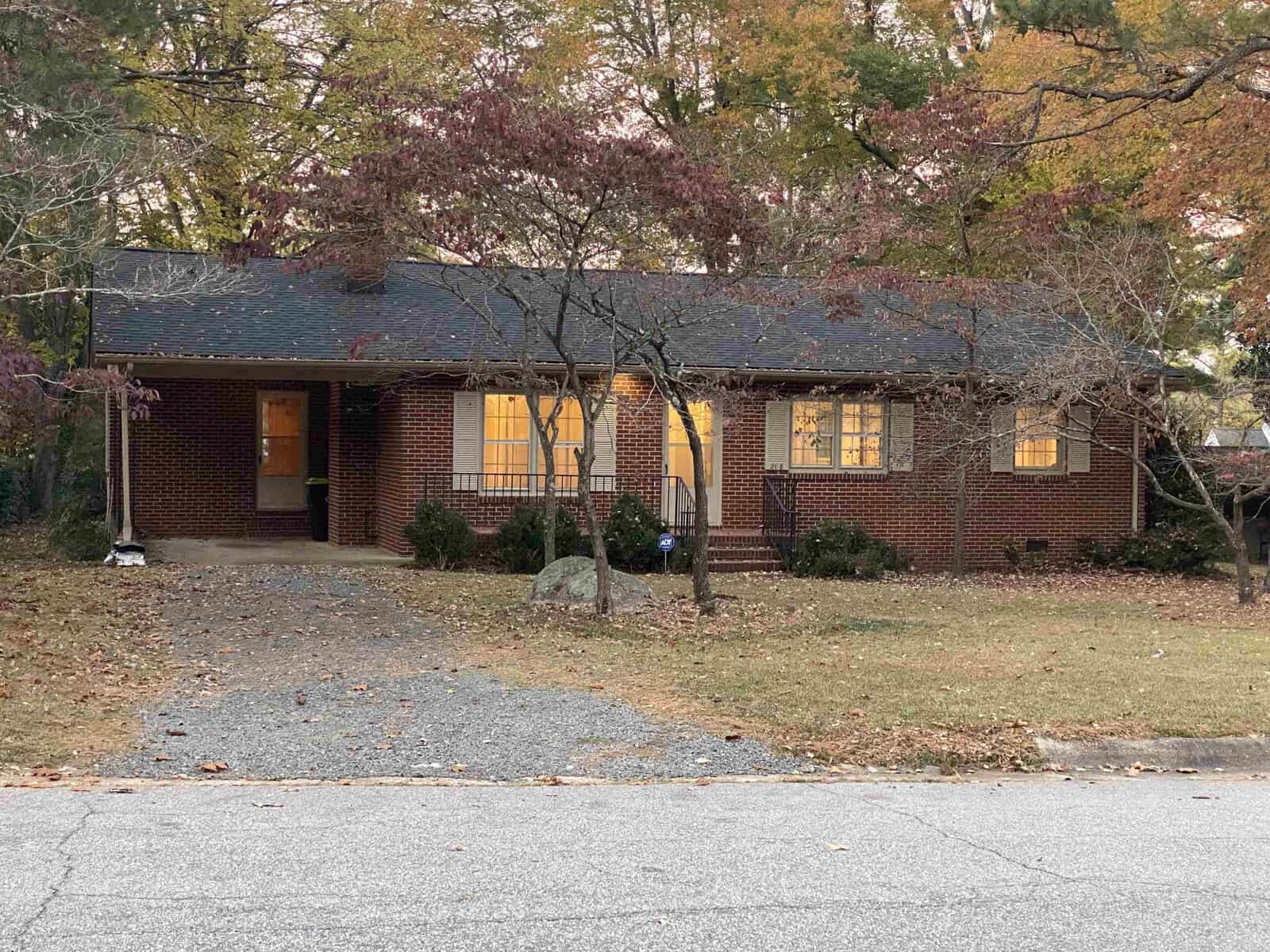 A brick house at 208 Hillcrest Drive, Oxford, with a lit interior, is surrounded by trees with autumn foliage. The house features a paved driveway and a small front yard sprinkled with fallen leaves. Twilight adds a warm glow to this picturesque scene.