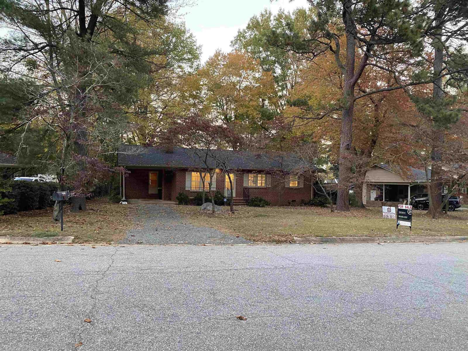 A single-story brick house with a dark roof is surrounded by tall trees with autumn leaves. The lawn is spacious, and a "For Sale" sign is visible in front of the house. The driveway is empty, and the sky is overcast.