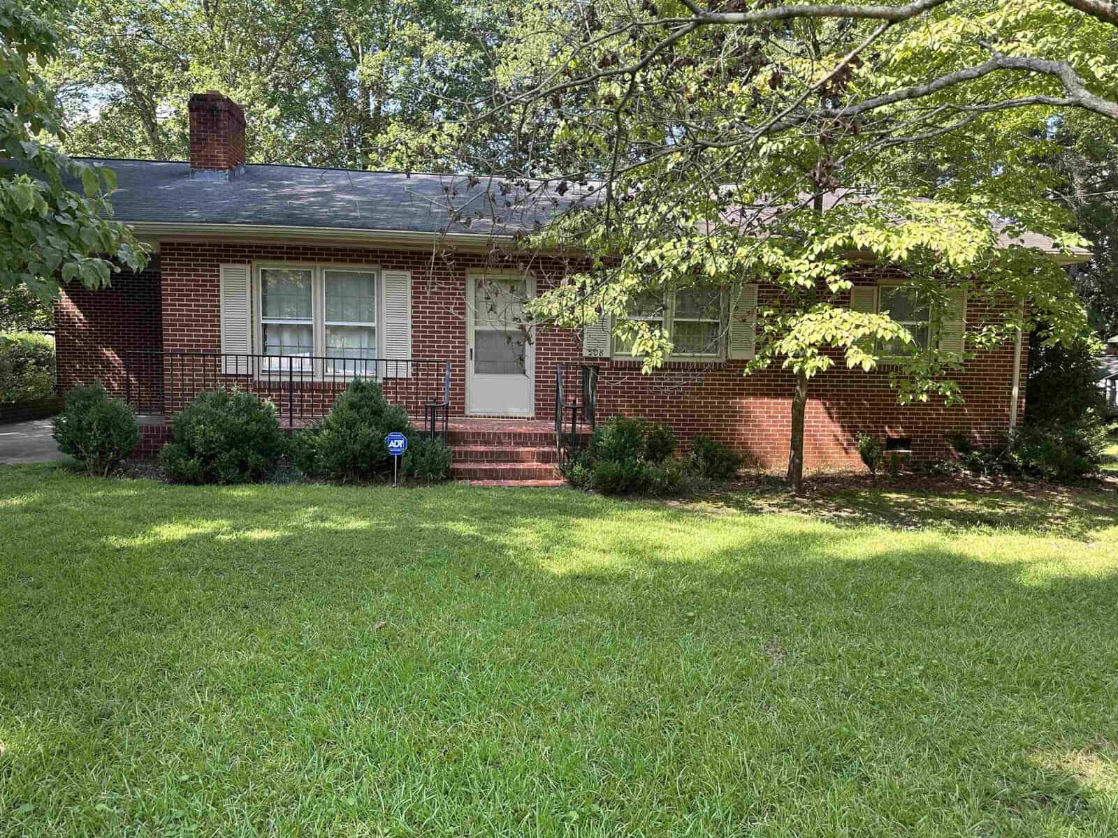 A brick, single-story house with a dark roof sits behind a lawn on a sunny day. A pathway leads to the front door, flanked by bushes. Trees provide shade and surround the property.