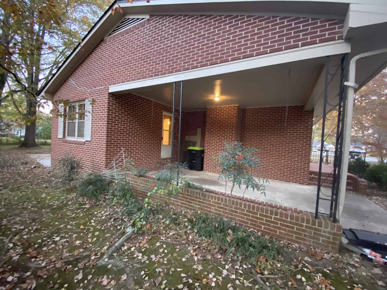 A brick house with a covered back porch showing outdoor lighting, a garbage bin, and potted plants. The surrounding area has grass and fallen autumn leaves.