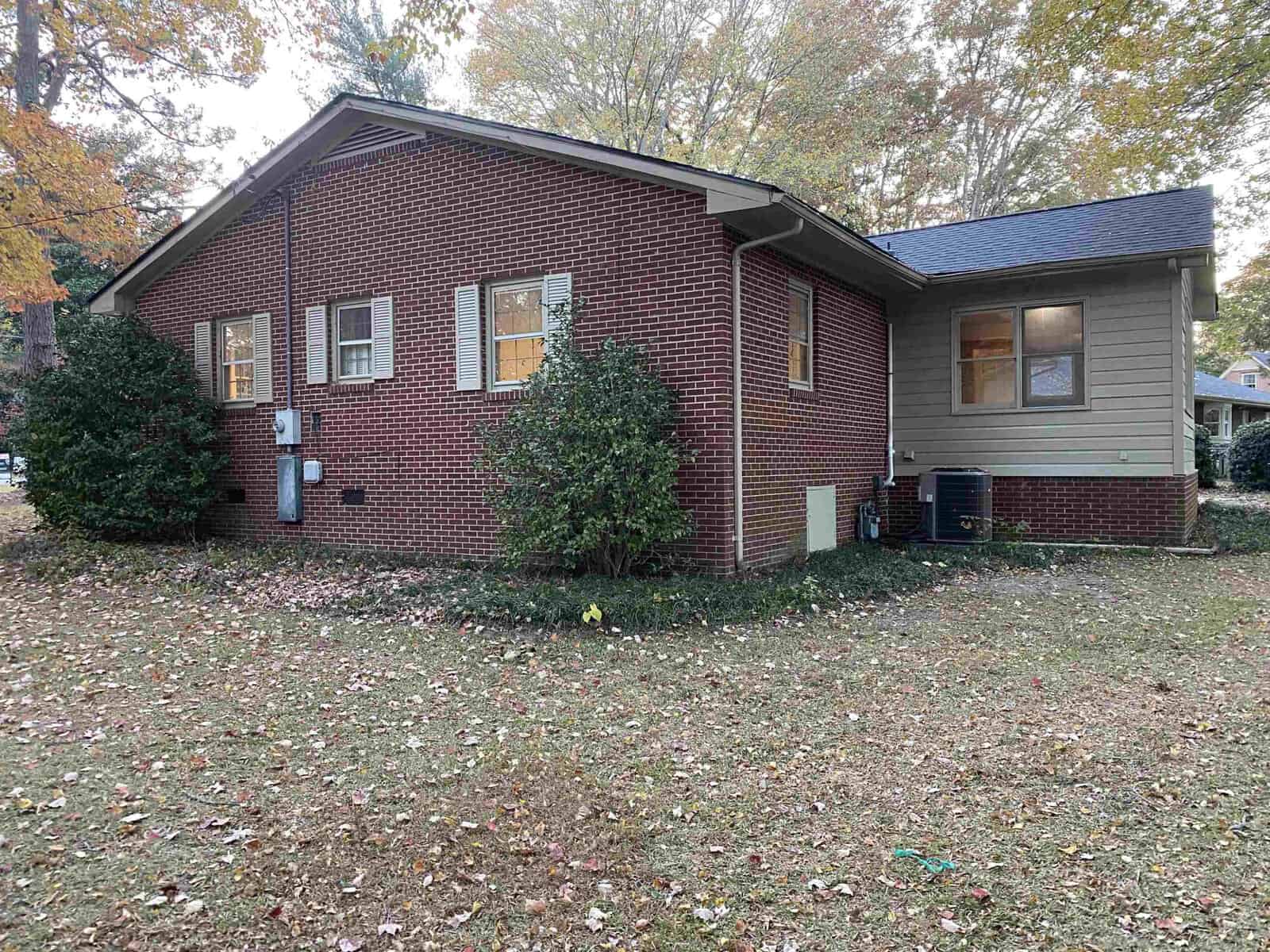 A brick and siding house with a dark roof, surrounded by a grassy yard and autumn trees. Several windows are visible, along with some bushes near the building. Fallen leaves cover parts of the ground.