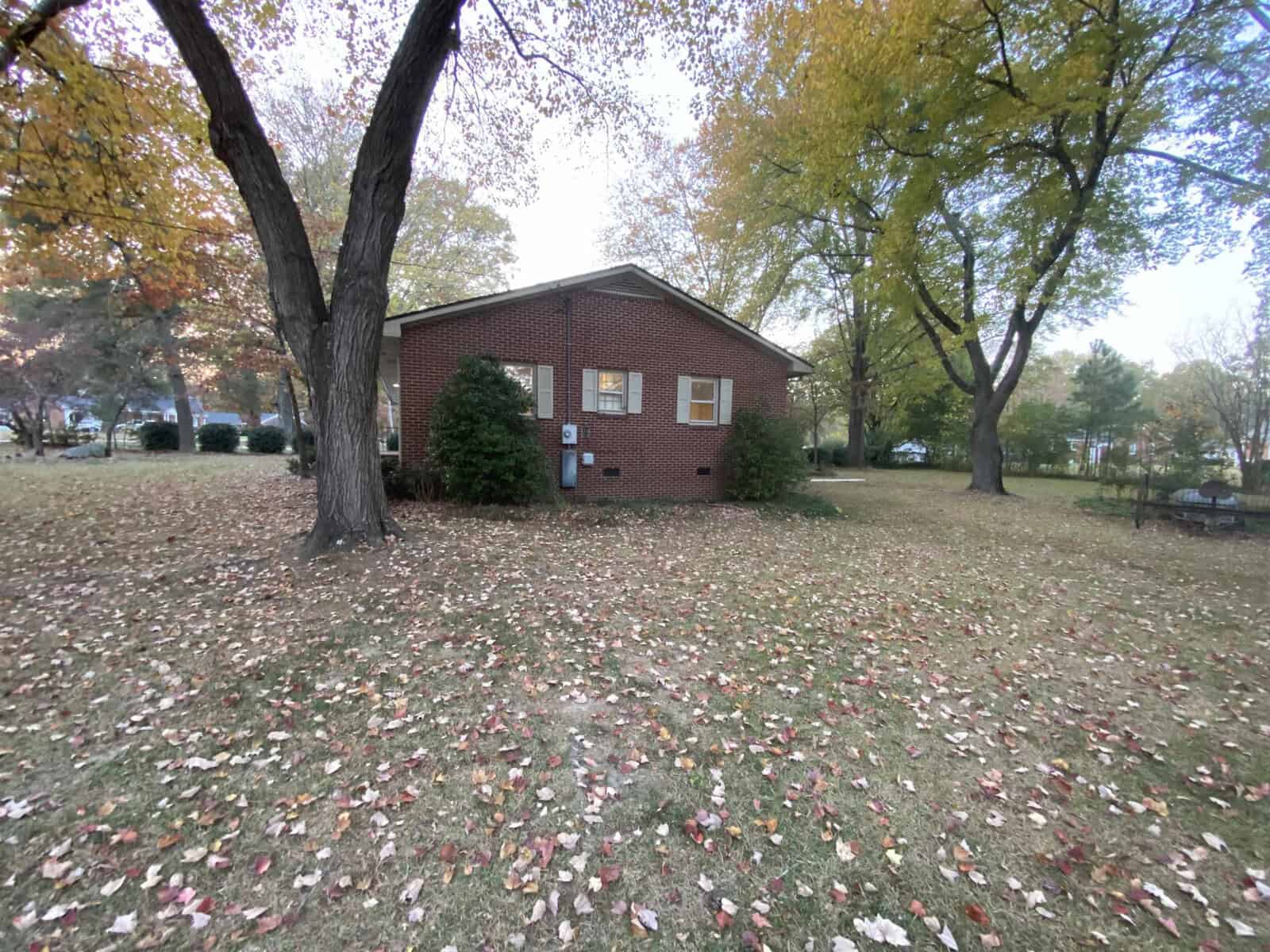 A small brick house with yellow shutters sits amidst a yard covered in autumn leaves. Large trees with yellow and green foliage surround the house, and the sky appears overcast.