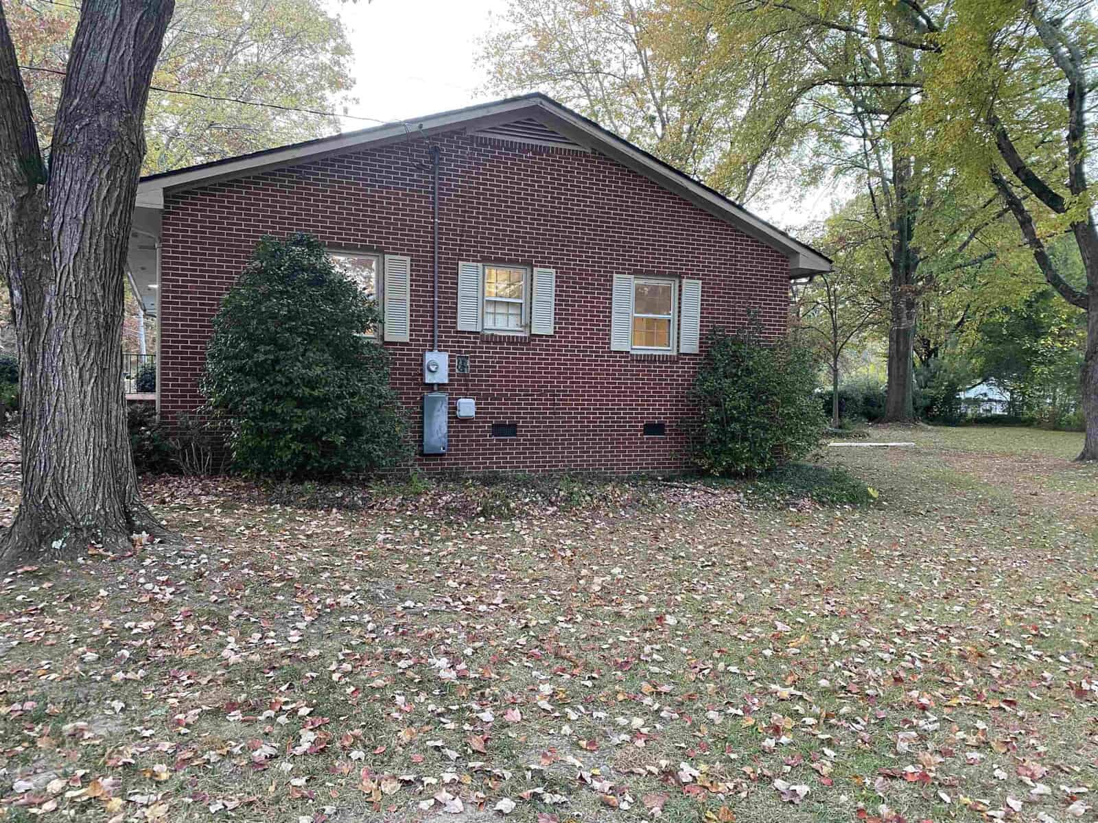 A brick house with large windows is surrounded by green bushes and trees. The lawn is covered in fallen leaves, indicating autumn. The house has a sloping roof, and there is a tree trunk in the foreground.