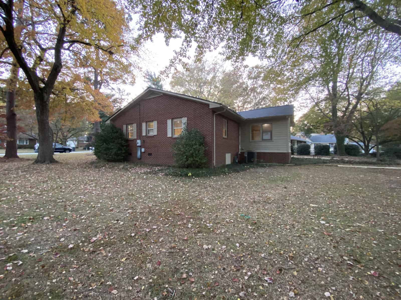 A single-story brick house with a large, leaf-covered lawn is surrounded by trees in autumn. The house has multiple windows and a small side extension. The sky is overcast, creating a serene, quiet atmosphere.