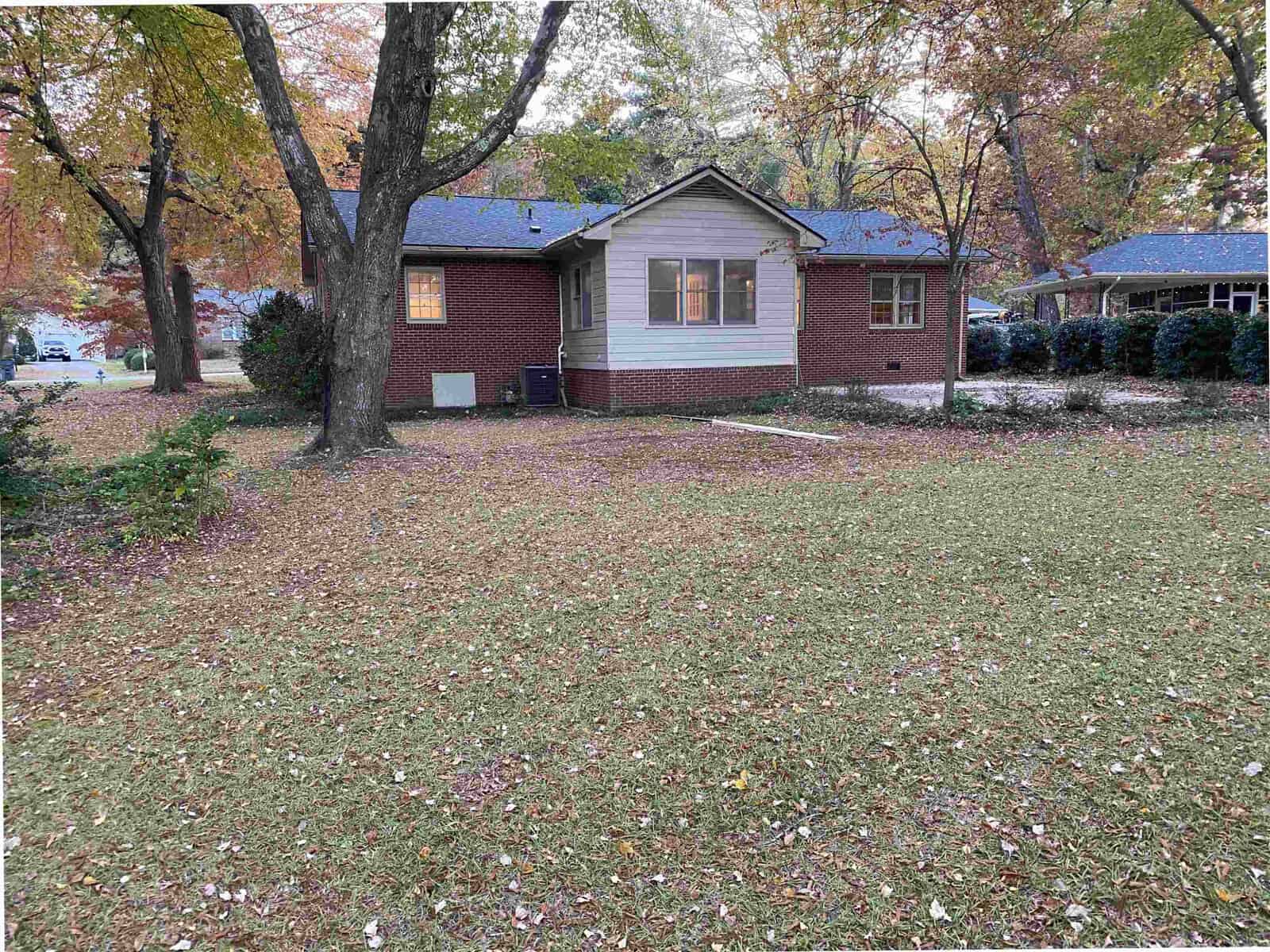 A brick house with a pitched roof is surrounded by autumn trees. The lawn is covered with fallen leaves, and there are two large trees in the foreground. The house has light-colored siding on the front section.