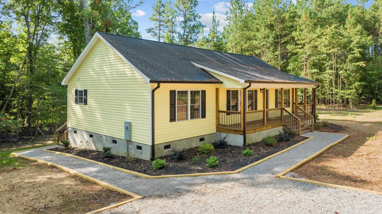 A single-story yellow house with a gabled roof and front porch, surrounded by a forested area with a concrete pathway leading to the entrance.
