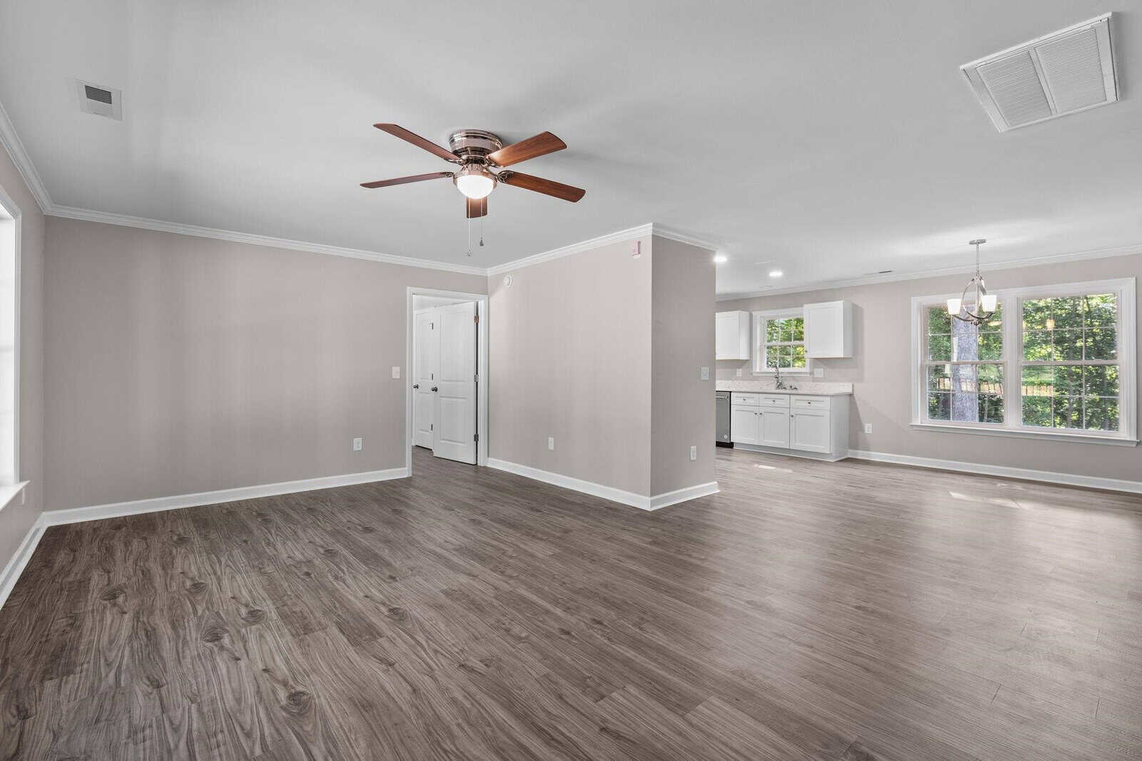 Empty modern living space with hardwood floors, white walls, ceiling fan, and view into a kitchen with white cabinets.