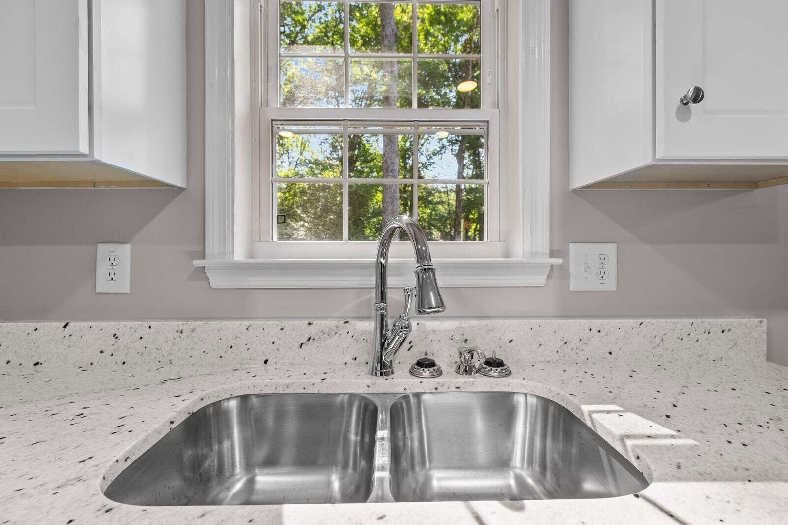 A double basin stainless steel sink installed in a kitchen countertop with a window above displaying a view of greenery outside.