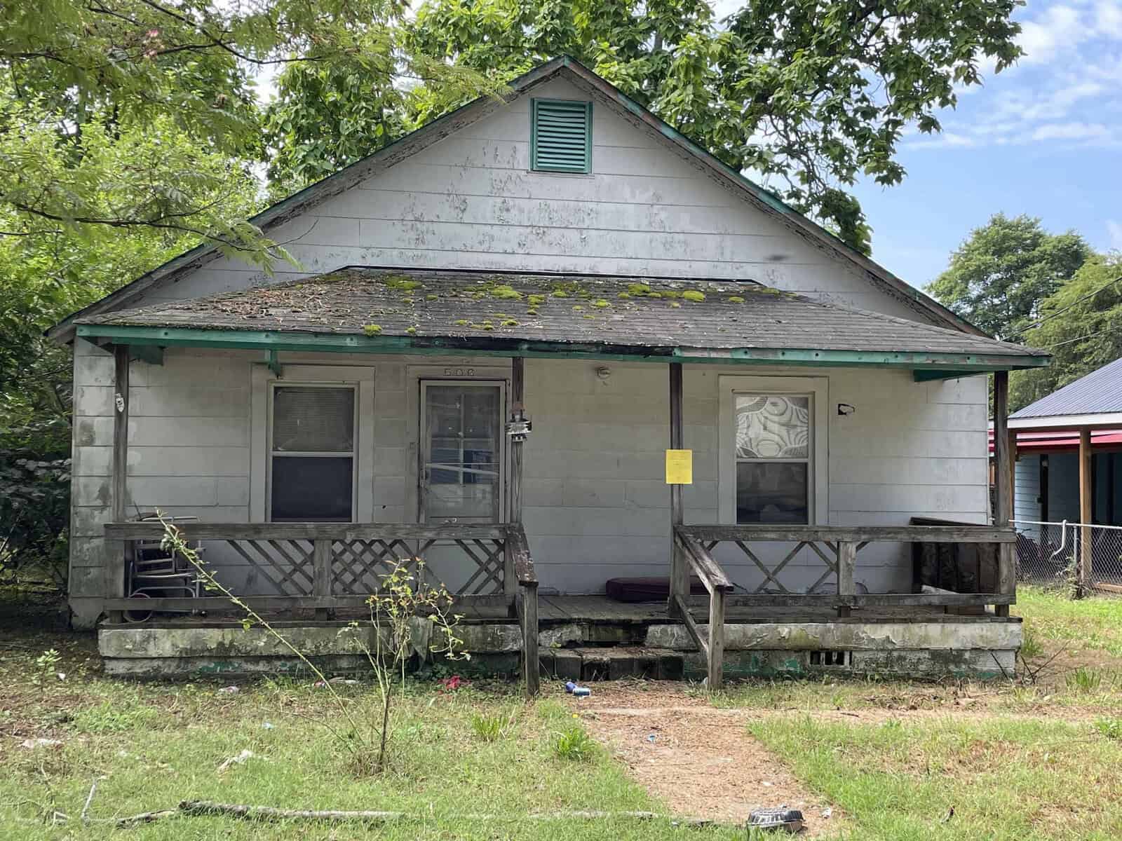 A weathered single-story house at 506 Hickory Street, Henderson NC, with a front porch, featuring peeling paint and a yellow notice attached to the front, set against a backdrop of over