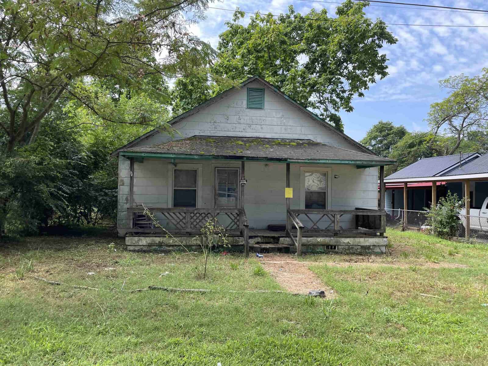 A small, weathered house with a front porch and overgrown yard, surrounded by greenery.