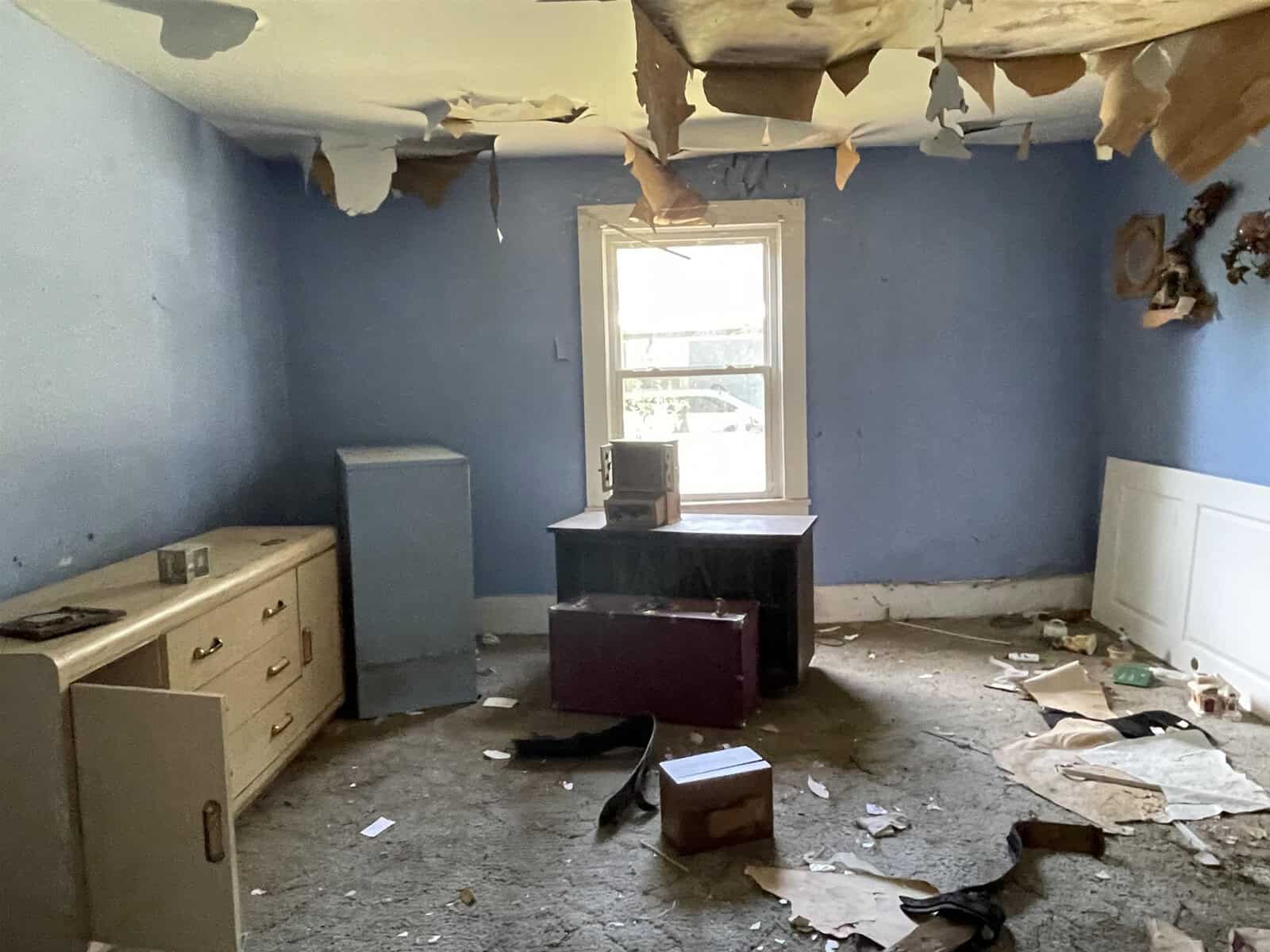 An abandoned room with peeling ceiling paint, scattered debris, and decayed furniture, bathed in natural light from a window.
