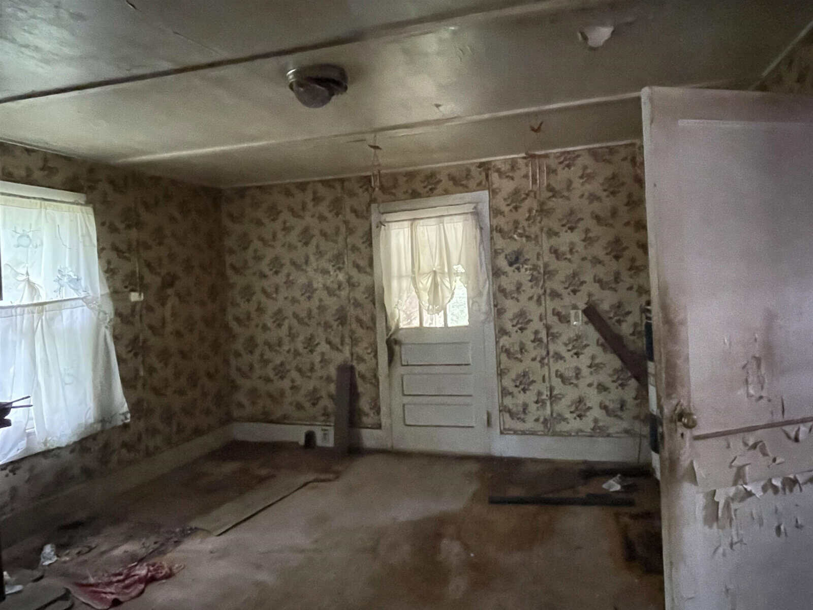 An abandoned room with peeling floral wallpaper, a dirty bare floor, and a door with a window adorned by a sheer curtain.