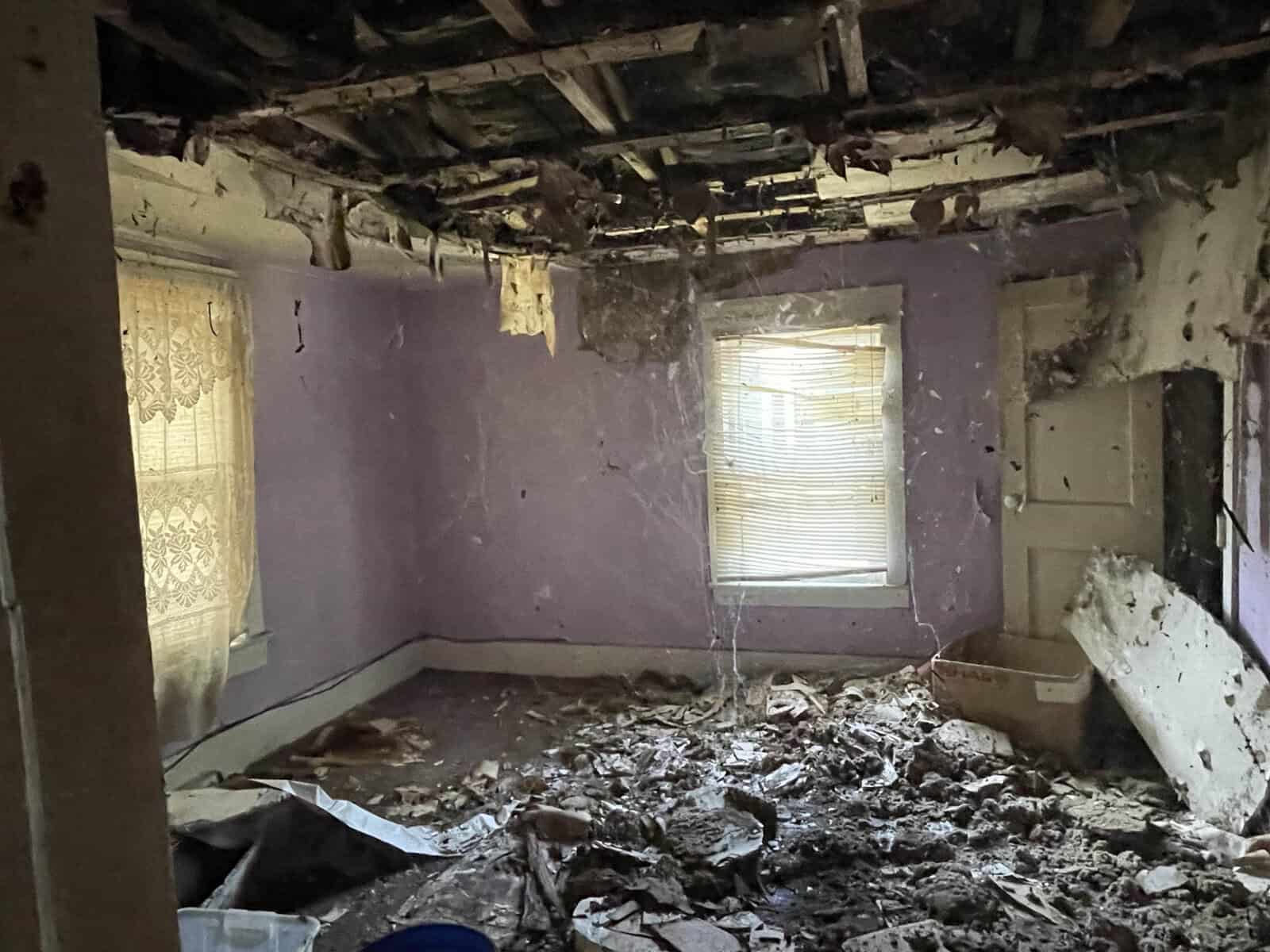 An interior room with significant damage and debris, featuring a collapsed ceiling, exposed beams, and destruction on the floor, likely due to an incident or ongoing decay.