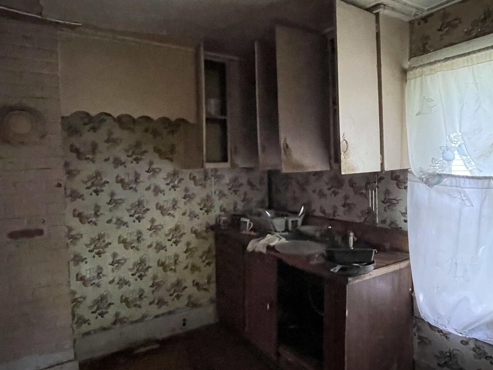 An old, neglected kitchen with dated wallpaper, open cabinets, and a dirty sink full of dishes.