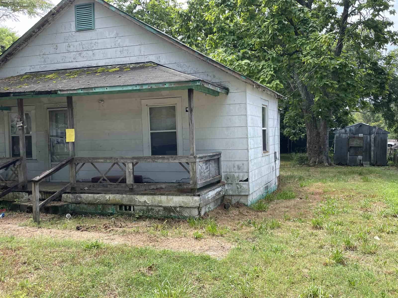A weathered single-story house with a dilapidated wooden porch and a notice posted on the front, surrounded by an unkempt yard with overgrown grass.