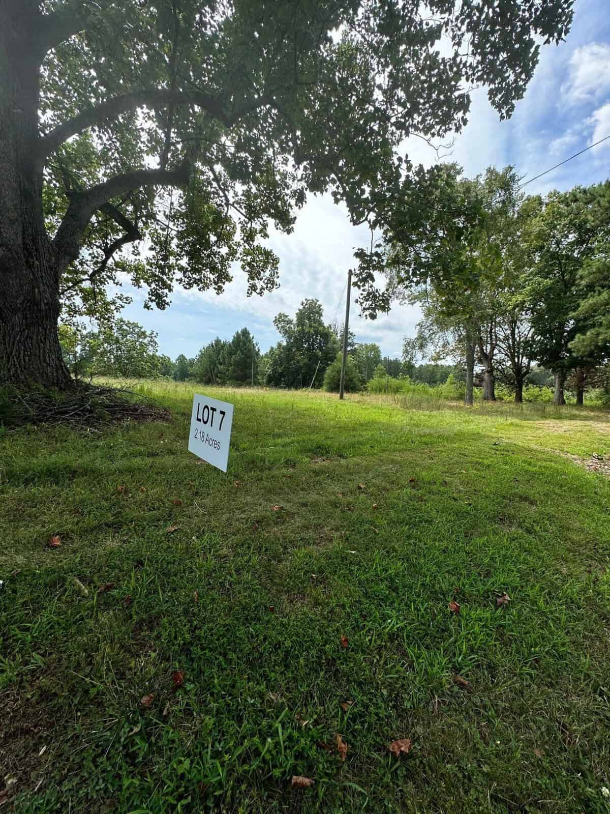 Grassy area with scattered trees under a partly cloudy sky, located at 3226 Bruce Garner Road. A small sign on the ground reads "LOT 7, 2.36 Acres." Nearby are utility poles and a dirt path, adding to the rustic charm of Creedmoor.
