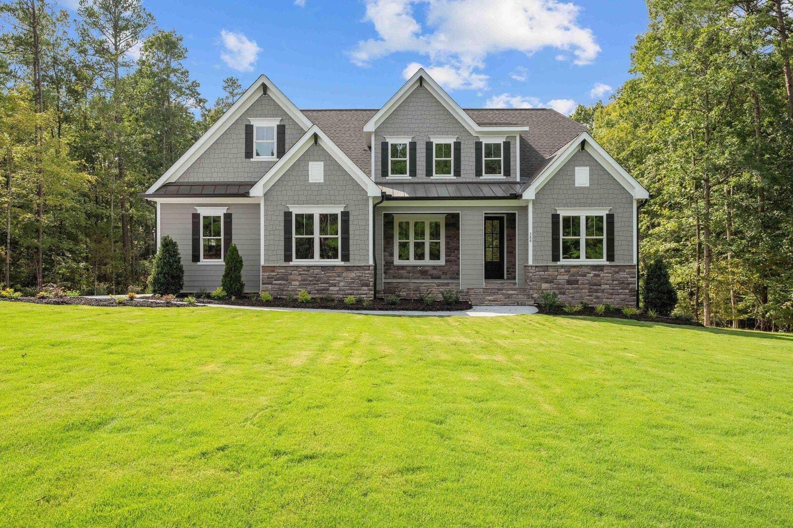 A two-story suburban home with a gray exterior and stone accents, surrounded by a freshly mowed lawn and set against a backdrop of trees under a clear sky.