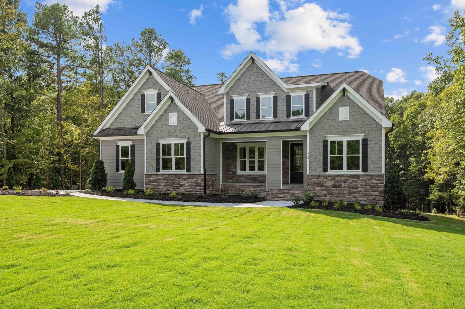A modern two-story house with gray siding, stone accents, a covered porch, and a well-manicured front lawn surrounded by trees under a clear sky.
