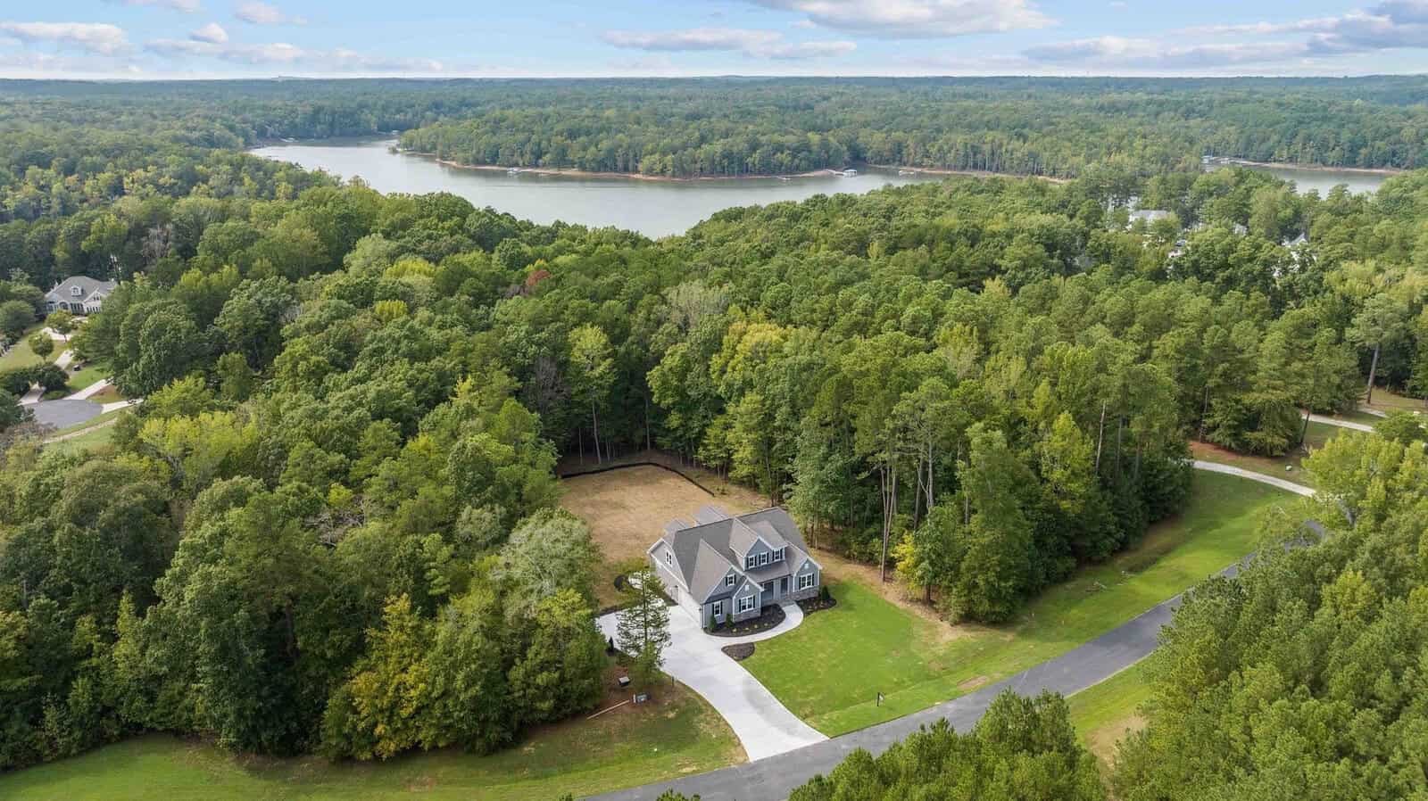 Aerial view of a solitary house surrounded by dense greenery near a large lake with residential areas in the distance.