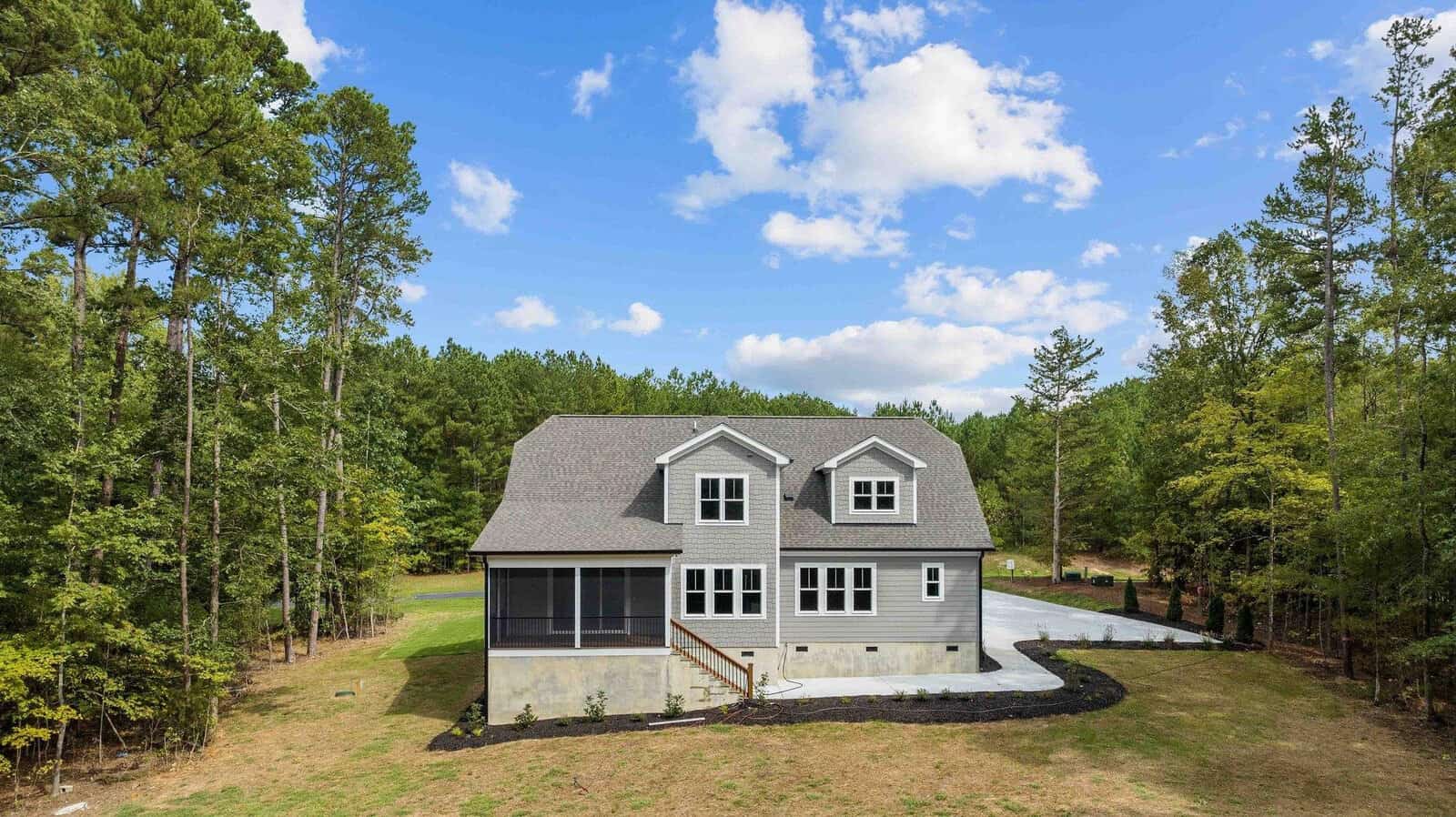 A two-story house with a dark gray roof and light-colored siding, featuring a large screened porch, situated in a clearing surrounded by tall pine trees under a partly cloudy sky. there is a newly paved driveway on the right side of the house.